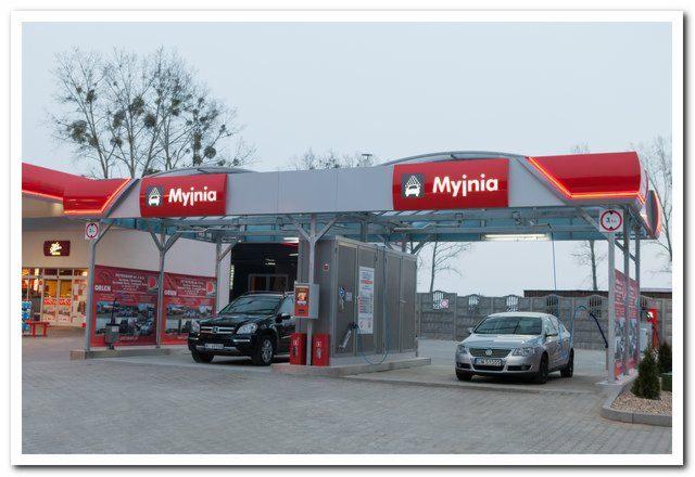 Stacja samoobsługowej myjni samochodowej Myjnia z dwoma stanowiskami, czarnym SUV-em i srebrnym sedanem, z widocznymi automatami do płatności i reklamami na ścianach.