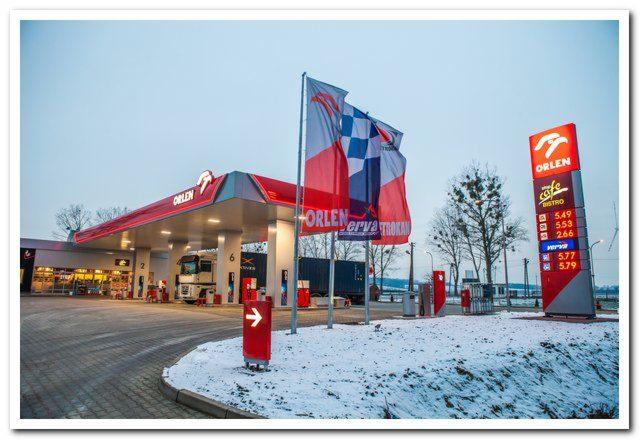 Stacja paliw Orlen z zaparkowaną ciężarówką, widoczna cena paliwa na pylonie i flaga reklamowa na tle lekko ośnieżonego terenu.