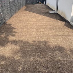 OG-ogrody - Wyrównany teren pod trawnik, pokryty nasionami trawy, otoczony ciemnoszarym drewnianym płotem. W tle nowoczesne budynki i jednostka klimatyzacji na ścianie.
