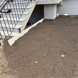 MTK Mateusz Tomasz Kuźniar - Świeżo przygotowana ziemia pod trawnik lub nasadzenia, oddzielona betonowym krawężnikiem od granitowych schodów z kutą balustradą przy szarej elewacji budynku.