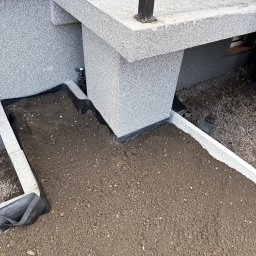 MTK Mateusz Tomasz Kuźniar - Przygotowany teren pod nasadzenia przy fundamencie budynku z szarym tynkiem mozaikowym. Widoczna warstwa ziemi, betonowe obrzeża i czarna folia, oddzielające od żwirowego podłoża.