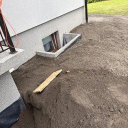 MTK Mateusz Tomasz Kuźniar - Świeżo nawieziona ziemia wokół ściany domu z oknem piwnicznym i betonową studzienką. Widoczna rura spustowa, schody i teren po niwelacji.