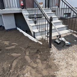 MTK Mateusz Tomasz Kuźniar - Wejście do domu z szarymi granitowymi schodami i czarnymi, kutymi balustradami. Obok schodów świeżo rozłożona ziemia z widocznymi śladami stóp, przechodząca w żwir, z narzędziami budowlanymi...