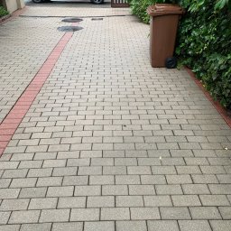 VaskPro - Czysta, szara kostka brukowa na podjeździe z czerwonym pasem, obok brązowy kosz na śmieci i zielony bluszcz. W tle zaparkowany ciemnoszary samochód. Brak mchu i zabrudzeń.