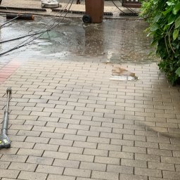 VaskPro - Mokra, świeżo wyczyszczona nawierzchnia z jasnej kostki brukowej, z widoczną lancą myjki ciśnieniowej i okrągłym narzędziem do czyszczenia. W tle ciemny samochód i zielone krzewy.