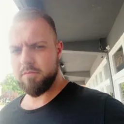 Mężczyzna z brodą w czarnej koszulce na tle budynku z oknami. Widoczne fragmenty elewacji i chodnika. Ujęcie od dołu, oświetlenie dzienne.