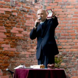 Mężczyzna w garniturze z mikrofonem prezentuje w pudełku medal na tle ceglanej ściany, obok stół z czerwonym obrusem i drewniany młotek.