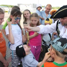 Mężczyzna przebrany za pirata karmi spaghetti grupę dzieci podczas zabawy na świeżym powietrzu, jedno dziecko ma pomalowaną twarz w motyle.