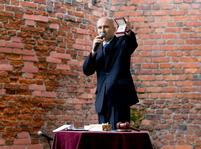 Mężczyzna w garniturze z mikrofonem prezentuje w pudełku medal na tle ceglanej ściany, obok stół z czerwonym obrusem i drewniany młotek.