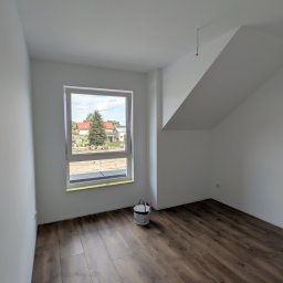 PRIMEWALL malowanie i tapetowanie wnętrz - Puste, świeżo pomalowane białe pomieszczenie z drewnianą podłogą, dużym oknem z widokiem na zielone drzewo i domy, oraz wiadrem farby. Widoczny skośny sufit.
