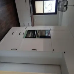 Biała zabudowa kuchenna z piekarnikiem w nowoczesnym stylu. Uwagę zwracają czarne, podłużne uchwyty i drewniana podłoga w ciepłym odcieniu brązu.