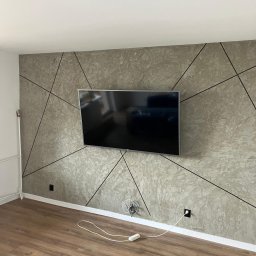 Sebastian Bielski - Ściana w salonie z telewizorem, wykończona szarym tynkiem dekoracyjnym o nieregularnej fakturze, z czarnymi, ostrymi liniami tworzącymi dynamiczny, geometryczny wzór. Widoczne panele podłogowe.