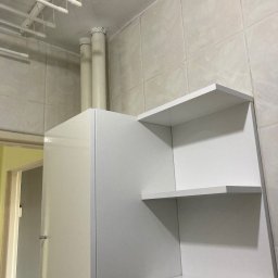 Hanna Mykhailova - Biała szafka z połyskiem i otwarte półki w narożniku pomieszczenia gospodarczego, z sufitową suszarką na pranie i beżowymi płytkami ściennymi. Widoczne rury instalacyjne.