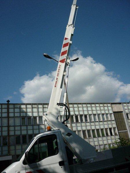 Podnośnik koszowy 18m na białym samochodzie dostawczym, wysięgnik z czerwono-białymi pasami ustawiony pod kątem, w tle budynek z licznymi oknami i błękitne niebo z chmurami.