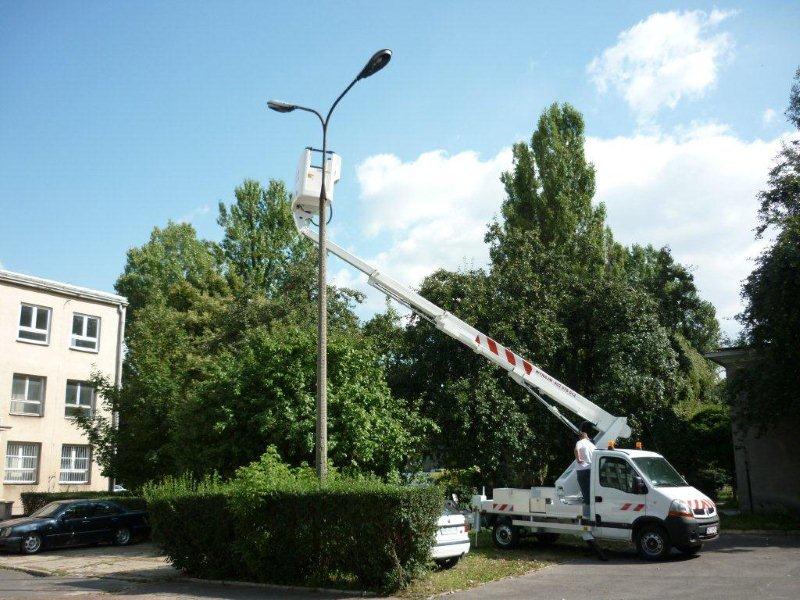 Podnośnik koszowy na tle budynku i drzew, skierowany w stronę słupa oświetleniowego, z koszem na wysokości lampy. Widoczny fragment samochodu osobowego i żywopłot.