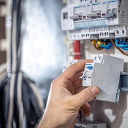 Zakład Usług Elektrycznych Jarosław Moliński - Męska dłoń instalująca szary wyłącznik nadprądowy na szynie DIN w otwartej rozdzielnicy elektrycznej, z uporządkowanymi kolorowymi przewodami.