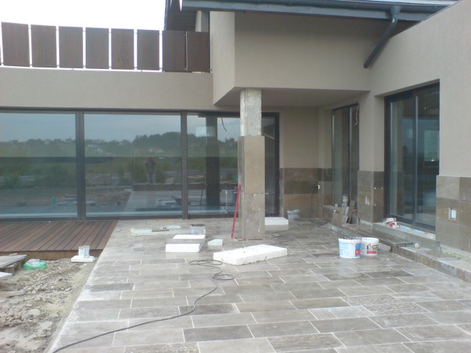 Układanie kamiennej posadzki na tarasie zewnętrznym nowoczesnego domu, widoczne płyty kamienne, materiały budowlane i narzędzia pracy, zarys budynku w tle.