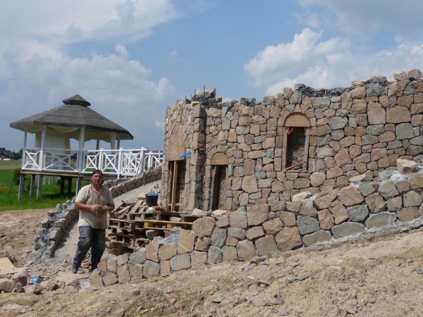 Budowa kamiennej konstrukcji z widocznym pracownikiem i altaną w tle, ukazująca etap wznoszenia ścian z kamienia naturalnego z przygotowanymi otworami okiennymi i drzwiowymi.