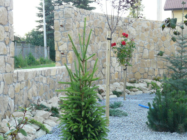 Kamienne ogrodzenie z jasnego piaskowca, fragment ogrodu z młodymi drzewkami, krzewami iglastymi i wysypanym grysem, widoczne elementy konstrukcyjne ogrodzenia.