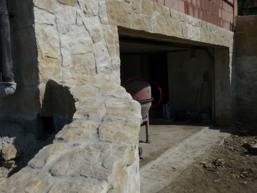 Wykończenie kamieniem elewacji garażu z widoczną betoniarką w środku, fragment murku z kamienia na pierwszym planie.