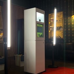 FotobudkaPlus - Nowoczesna, biało-szara budka do zdjęć z ekranem, oświetlona pionowymi lampami LED, na czerwonym dywanie w eleganckim wnętrzu z ciemnozieloną tapetą w złote wzory.