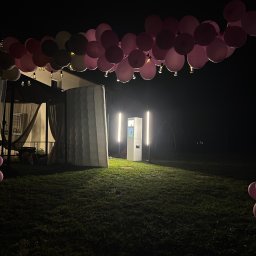 FotobudkaPlus - Elegancki, biały kiosk fotograficzny z pionowym oświetleniem LED, stojący na nocnym trawniku pod łukiem różowych balonów i girland świetlnych.