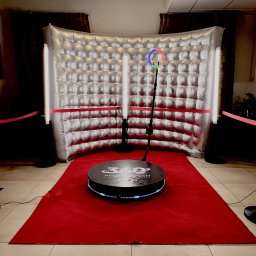 FotobudkaPlus - Ustawiona fotobudka 360 z czarną platformą na czerwonym dywanie, otoczona srebrną, pikowaną ścianką i pionowymi lampami LED. Widoczny kod QR do skanowania.