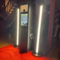 FotobudkaPlus - Czarne urządzenie do zdjęć z ekranem dotykowym i dwoma pionowymi lampami LED, ustawione na czerwonym dywanie. W tle drewniane schody, balustrada i dekoracje eventowe.