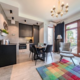 Ostrowska Nieruchomości - Nowoczesny apartament z aneksem kuchennym w antracycie, okrągłym stołem jadalnym i fotelem obok dużego okna balkonowego. Kolorowy dywan ożywia przestrzeń.