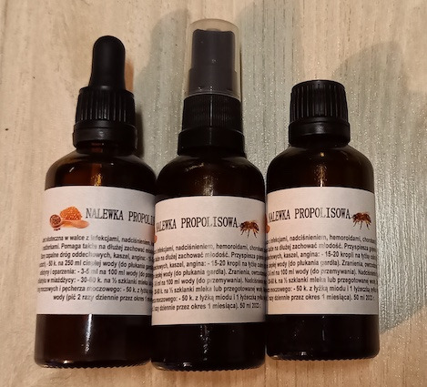 Trzy brązowe butelki z etykietami 'Nalewka Propolisowa' i 'Nalewka Propolisowa' z różnymi aplikatorami: kroplomierz, atomizer i zakrętka, ustawione blisko siebie na jasnym tle.