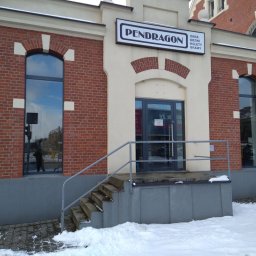 PENDRAGON - Fasada budynku z cegły i beżowego tynku, z ciemnymi oknami łukowymi i prostokątnymi oraz przeszklonymi drzwiami. Szyld Pendragon reklamuje okna, drzwi, rolety, bramy. Przed wejściem schody i śnieg.