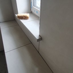 Usługi Remontowo-Budowlane - Jasne, matowe płytki na ścianach ze skosami i szerokim parapecie wokół okna PCV z folią ochronną. Ciemne fugi i gąbka na parapecie wskazują na trwające prace.
