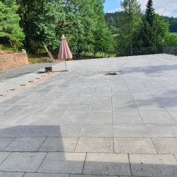 Usługi Remontowo-Budowlane - Rozległy taras z szarych płyt w trakcie układania, z widoczną piłą do cięcia, zamkniętym parasolem i ozdobną balustradą. W tle zielony krajobraz.