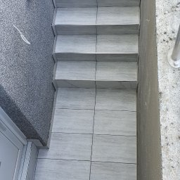 Usługi Remontowo-Budowlane - Ukończone schody zewnętrzne i podest wyłożone jasnoszarymi płytkami z metalowymi listwami, obok ściana z tynku mozaikowego i kratka odpływowa.
