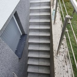 Usługi Remontowo-Budowlane - Szare, kafelkowane schody zewnętrzne prowadzące do piwnicy, obok ściana z dwoma rodzajami tynku i okno z roletą. Stalowa balustrada i trawnik.
