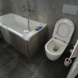Usługi Remontowo-Budowlane - Nowoczesna łazienka z ciemnoszarymi płytkami na podłodze i ścianach, białą wanną z chromowaną baterią oraz podwieszanym WC. Na parapecie okna stoją dwie bursztynowe butelki.