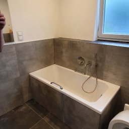 Usługi Remontowo-Budowlane - Nowoczesna łazienka z białą wanną w zabudowie, wykończona dużymi, ciemnoszarymi płytkami na ścianach i podłodze, z oknem z matowego szkła i wentylatorem ściennym.