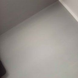 Mateusz Choczaj - Czysty narożnik pokoju z jasnoszarą podłogą, jedną ścianą w ciemniejszym szarym odcieniu i drugą białą z białym cokołem. Widoczny fragment ciemnego obiektu.