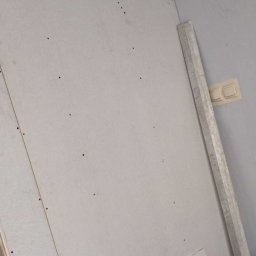 Mateusz Choczaj - Montaż suchej zabudowy na ścianie, z widocznymi wkrętami mocującymi i metalowym profilem opartym o narożnik, obok beżowy włącznik światła.