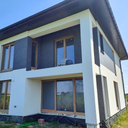 AUTOREMBUD SPÓŁKA Z OGRANICZONĄ ODPOWIEDZIALNOŚCIĄ - Dwukondygnacyjny dom z jasną elewacją i ciemnymi akcentami, widok z dołu, okna z drewnianymi ramami, balkon z krzesłami, błękitne niebo w tle.