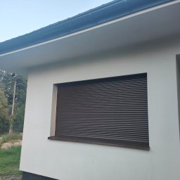 AUTOREMBUD SPÓŁKA Z OGRANICZONĄ ODPOWIEDZIALNOŚCIĄ - Biały budynek z brązowymi roletami zewnętrznymi na oknie. Widoczny fragment elewacji, okno z roletą i fragment dachu z rynną. Na zewnątrz trawa i drzewa.