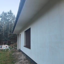 AUTOREMBUD SPÓŁKA Z OGRANICZONĄ ODPOWIEDZIALNOŚCIĄ - Ściana domu jednorodzinnego z brązową roletą okienną i widocznym fragmentem dachu. Na trawniku leżą resztki materiałów budowlanych. Widok z boku.