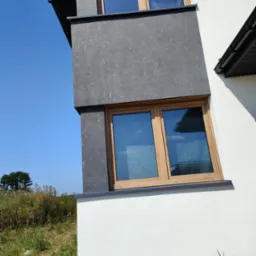 Fragment nowo wybudowanego domu z oknami w drewnianych ramach, szarą elewacją na górze i białą na dole, widok z dołu na tle błękitnego nieba.