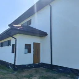 Nowo otynkowany, dwukondygnacyjny dom z ciemnym dachem i rynnami, widoczna folia fundamentowa oraz otwór okienny bez ramy, z częściowo zamontowanym ociepleniem.