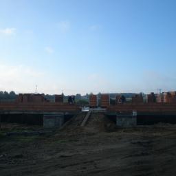 PIOTR ŚLADEWSKI ISO-GLAZ - Budowa domu w trakcie realizacji: widok na fundamenty, wznoszone ściany z czerwonej cegły i dwóch pracowników murujących, na pierwszym planie ziemia i przyczepka