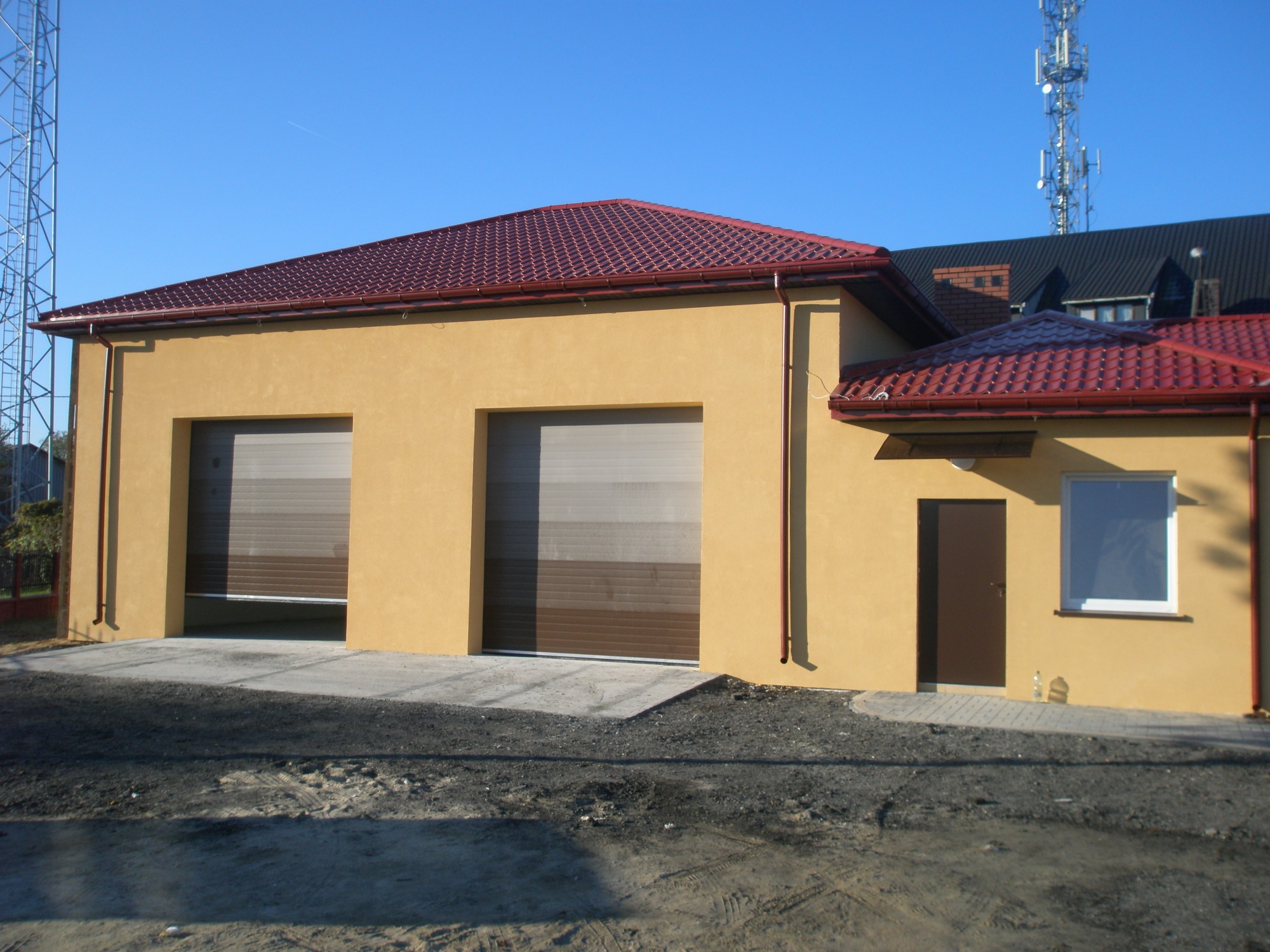Żółty budynek gospodarczy z dwoma bramami garażowymi i czerwoną dachówką na tle błękitnego nieba i wieży telekomunikacyjnej.
