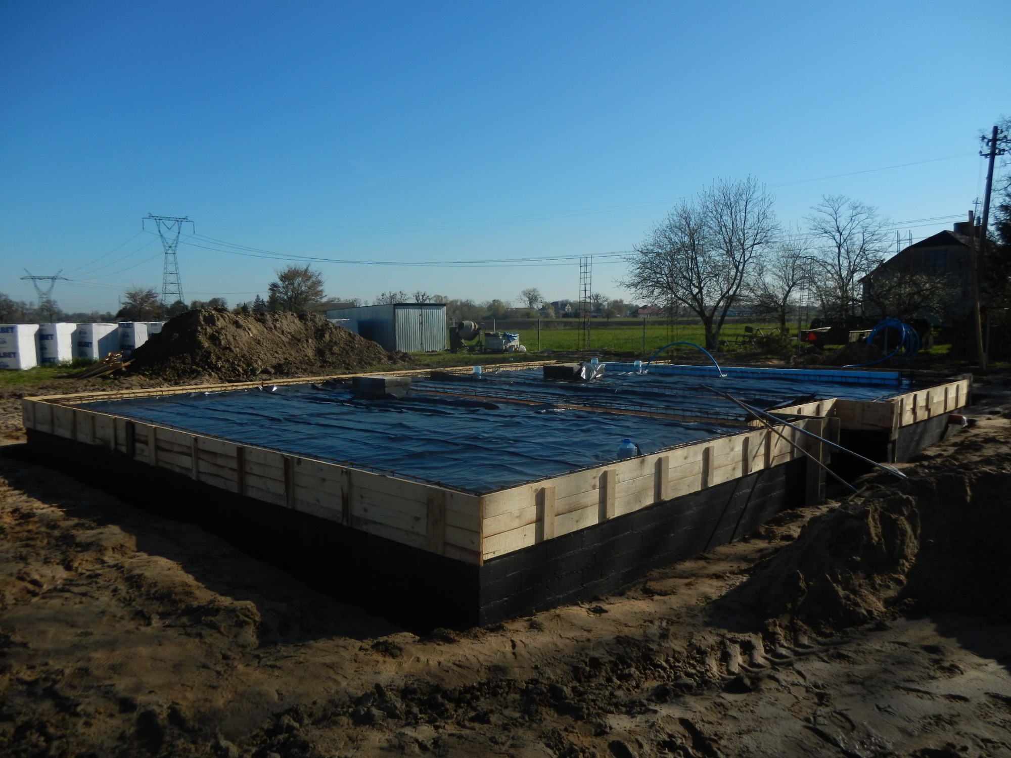 Przygotowanie fundamentów pod budowę domu: drewniany szalunek, izolacja przeciwwilgociowa, zbrojenie, widoczny wykop w ziemi.