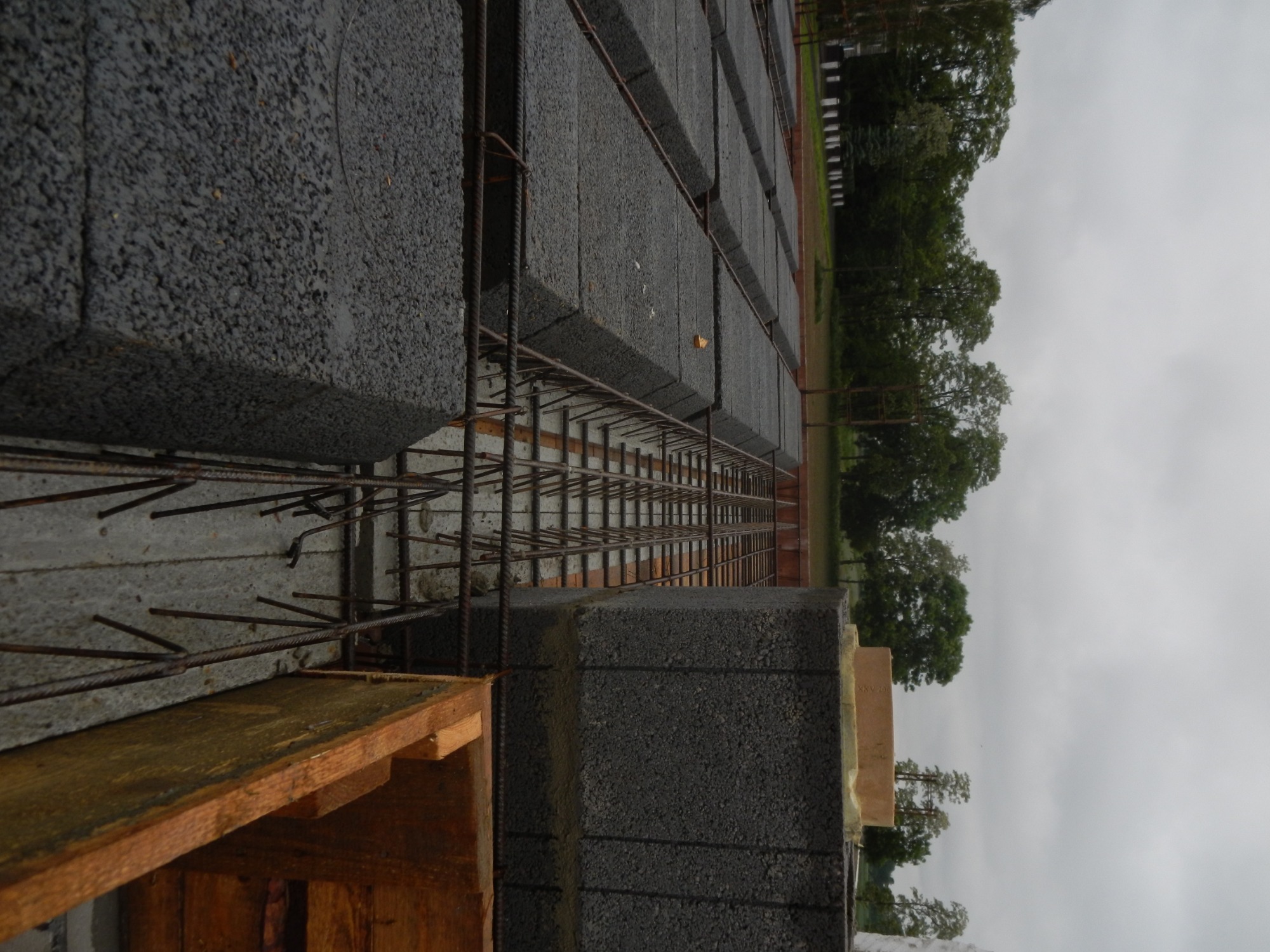 Wznoszenie ściany z szarych bloczków betonowych zbrojonych stalą na tle zieleni i pochmurnego nieba, widoczne drewniane elementy szalunku.