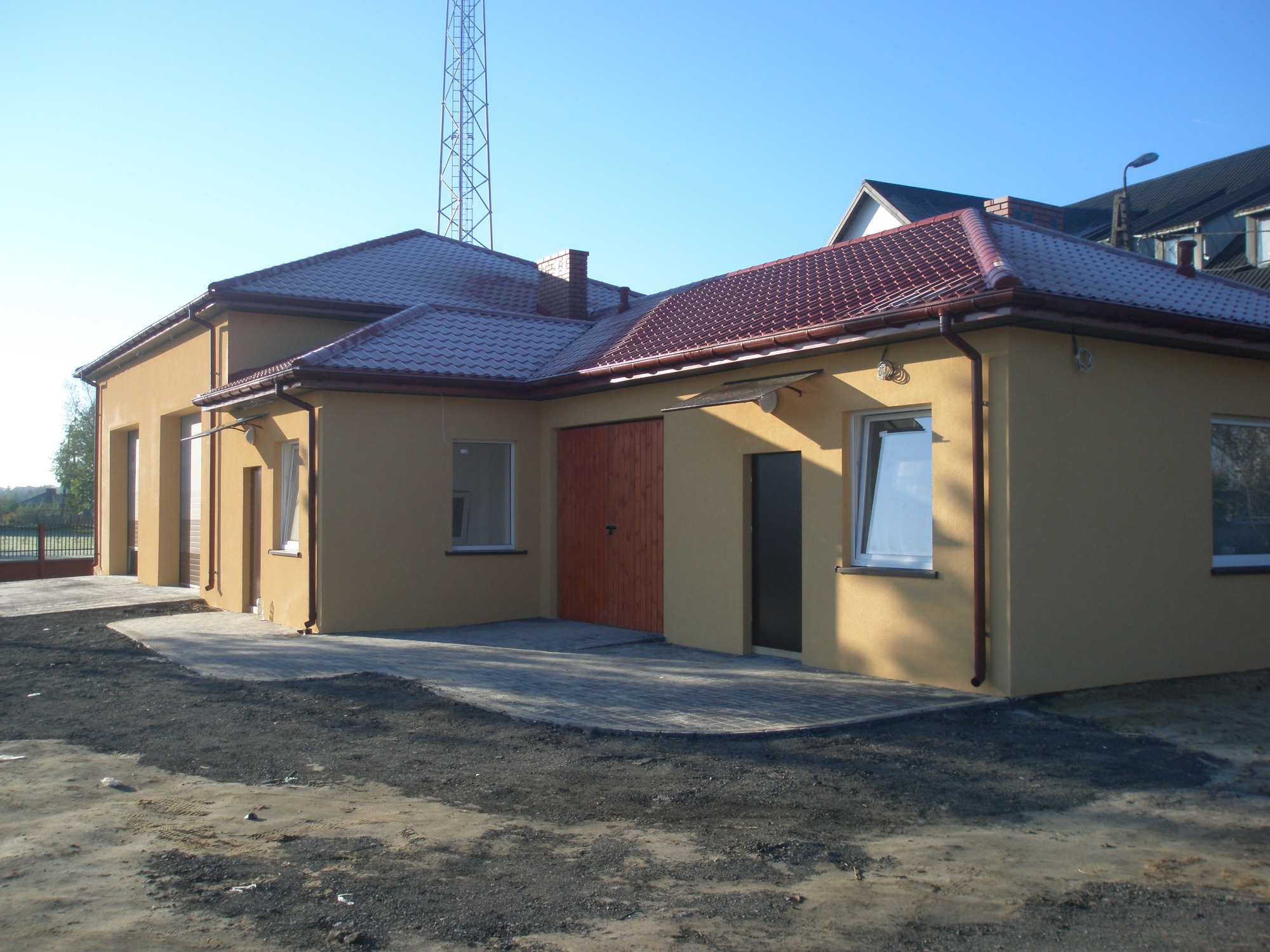 Parterowy dom murowany z czerwoną dachówką, świeżo otynkowany, z częściowo wybrukowanym podjazdem i widoczną konstrukcją masztu w tle.