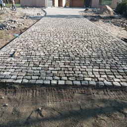 Katarzyna Zmiendak - Ułożony fragment podjazdu z nieregularnej szarej kostki granitowej, z widoczną podbudową piaskową i sznurkami poziomującymi, przed nowoczesnym domem w Krzęcinie.